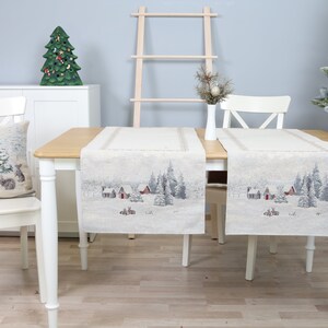 Può includere: Una scena festosa con due runner da tavolo e un cuscino su una sedia bianca. I runner e il cuscino presentano un paesaggio innevato con case e alberi. Un albero di Natale e decorazioni creano un'atmosfera festiva.