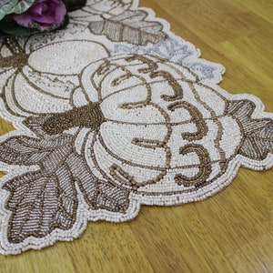 Può includere: Un runner da tavolo in perle bianche con un design di tre zucche e foglie. Le zucche sono contornate da perle marroni e le foglie da perle argentate.