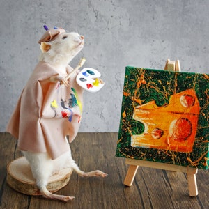 Może przedstawiać: Biały szczur w różowo-białej szacie i fioletowym kapeluszu trzyma pędzel i paletę farb. Szczur stoi na drewnianej platformie przed małym sztalugą z zielonym płótnem pomalowanym abstrakcyjnym wzorem w kolorze żółtym i pomarańczowym.