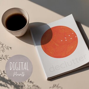 May include: A white book with an orange sun illustration and the word "declutter" is next to a cup of coffee. The book also has small white bird illustrations. The words "DIGITAL Prints" are in a gray circle.
