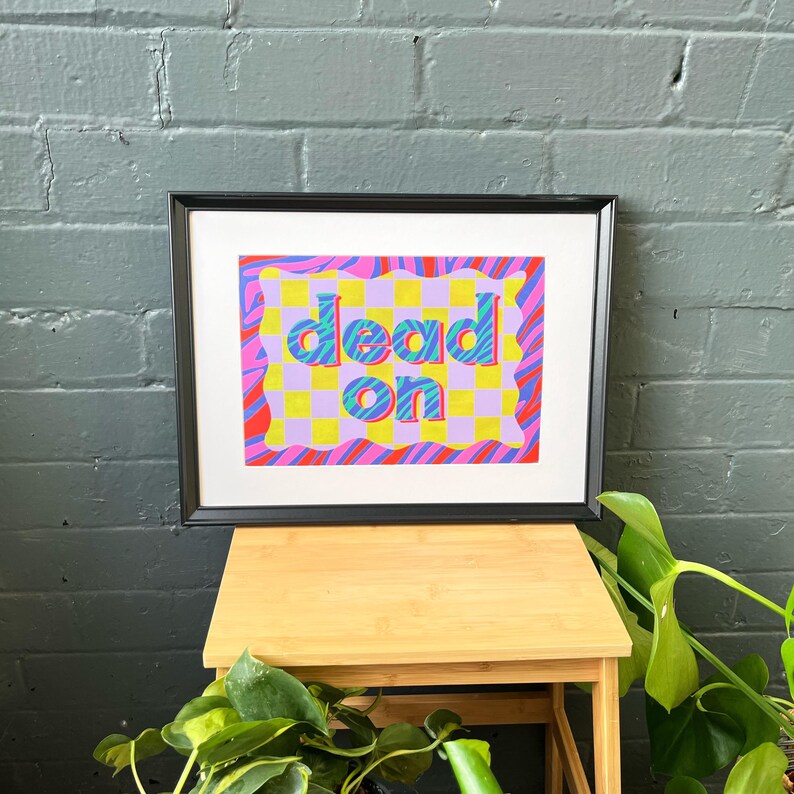 May include: A framed print with a colorful design featuring the words "dead on" in a bold, 3D-style font. The background is a checkered pattern with pink, yellow, and blue stripes.