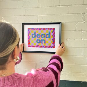 May include: A framed print with a colorful abstract design featuring the words "dead on" in a bold, blue font. The background is a mix of pink, yellow, and blue with a checkered pattern.