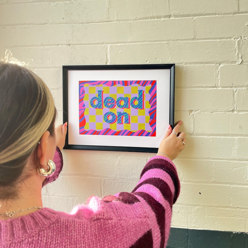 May include: A framed print with a colorful abstract design featuring the words "dead on" in a bold, blue font. The background is a mix of pink, yellow, and blue with a checkered pattern.