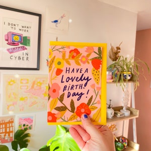 May include: A greeting card with a yellow border and a pink background, featuring colorful floral illustrations. The card reads "Have a Lovely Birthday!" in a handwritten style. The card is held in front of a white wall with framed art and a shelf with plants.