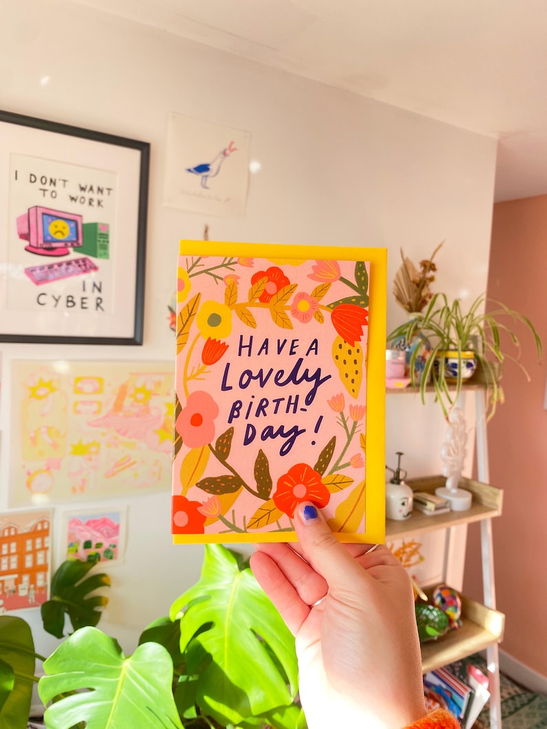 May include: A greeting card with a yellow border and a pink background, featuring colorful floral illustrations. The card reads "Have a Lovely Birthday!" in a handwritten style. The card is held in front of a white wall with framed art and a shelf with plants.