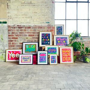 May include: A collection of framed prints with colorful, hand-drawn illustrations and text. The prints feature phrases like "Wind Yer Neck In", "Buck Efit", "Dead On", "That's Grand", "Aye", "Abs Make Ya Happy", "What's the Craic?", "Wee Dan Der", and "Yee".