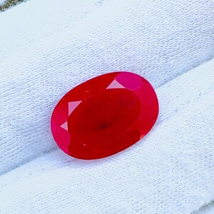 Peut inclure: Une pierre précieuse rouge vif, de forme ovale et à facettes. La gemme est posée sur un fond blanc, mettant en valeur sa couleur et sa taille. Les facettes de la pierre précieuse reflètent la lumière, créant un effet scintillant. Idéale pour la joaillerie.