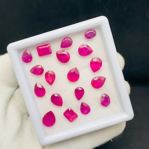 May include: A collection of 17 pink ruby gemstones in various shapes and sizes, displayed in a white box.