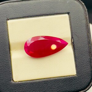 May include: A pear-shaped ruby gemstone with a red hue. The gemstone is set in a white background.
