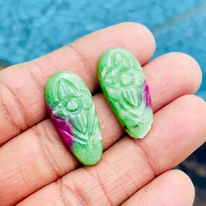 May include: Two green and pink carved gemstone cabochons with a floral design. The cabochons are oval shaped and have a smooth, polished finish.