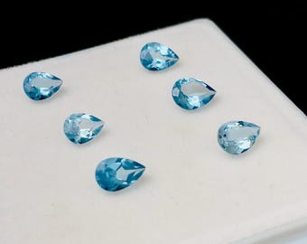 May include: Six pear-shaped blue topaz gemstones on a white surface.
