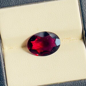 Peut inclure: Une pierre précieuse ovale, taillée à facettes, d'un rouge profond, sur un fond crème. La couleur de la pierre varie du foncé, presque noir, au rouge vif, avec la lumière se reflétant sur ses surfaces polies. Il s'agit probablement d'un grenat.