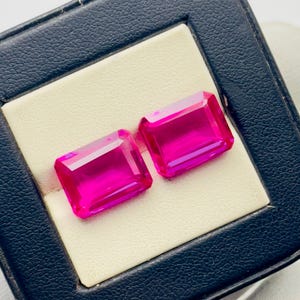 May include: Two vibrant pink emerald cut gemstones displayed in a black velvet jewellery box.