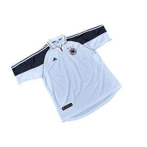 Op de afbeelding: Wit voetbaltenue met zwarte en grijze accenten. Het tenue heeft een ritskraag en het Adidas-logo op de linkerborst. Het embleem van het Duitse nationale team bevindt zich op de rechterborst.