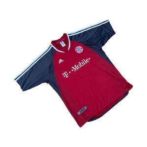 May include: A red and navy blue soccer jersey with white accents. The jersey features the Adidas logo, the Bayern Munich team emblem, and the "T-Mobile" sponsor logo. The sleeves are navy blue with white stripes.