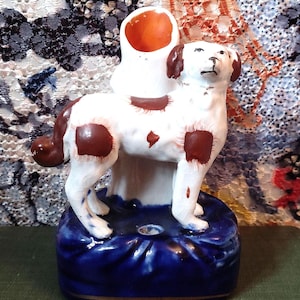 Könnte beinhalten: Eine weiße und braune Keramik-Hundefigur mit einem blauen und goldenen Sockel. Der Hund steht auf seinen Hinterbeinen und hat einen braunen Fleck auf dem Rücken. Die Figur ist im Vintage-Stil gehalten und hat einen rustikalen Look.