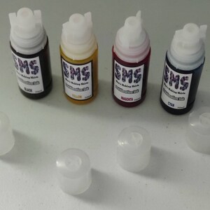 May include: Four small bottles of sublimation ink, labeled with the colors black, yellow, magenta, and cyan. The bottles are arranged in a row, with their caps removed and placed below them.