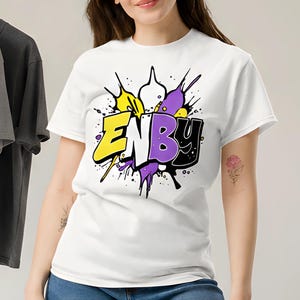 Può includere: Una maglietta bianca con la scritta "ENBY" in un design in stile graffiti, con accenti gialli, viola e neri. La modella indossa jeans blu. Ulteriori magliette bianche, grigie e nere sono appese a un attaccapanni sullo sfondo.