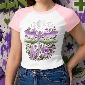 May include: A white and pink raglan crop top with a dragonfly design. The graphic features a dragonfly, crystals, and mushrooms. The background includes graffiti art with the words "Gender Queer", "Beyond Binary Beyond Limits", and "Not a Phase".