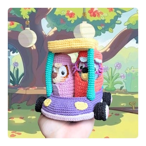 May include: A handmade crochet toy car featuring two animal characters. The car is purple with black wheels and a yellow roof. The characters are in pink and red, with teal accents. The background is a cartoon park scene.