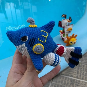 Könnte beinhalten: Ein blauer gehäkelter Hai mit weißen und roten Details. Der Hai hat eine gelbe Nummer "3" an der Seite und ein graues und gelbes kreisförmiges Detail am Kopf. Der Hai wird von einer Hand gehalten.