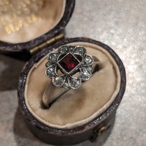 Pu&ograve; includere: Un anello color argento con una pietra preziosa rosso scuro a taglio quadrato al centro, circondata da un alone di dieci piccole pietre rotonde e trasparenti disposte a motivo floreale, presentato in un portagioie ovale marrone scuro foderato.