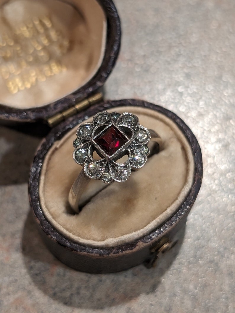 Pu&ograve; includere: Un anello color argento con una pietra preziosa rosso scuro a taglio quadrato al centro, circondata da un alone di dieci piccole pietre rotonde e trasparenti disposte a motivo floreale, presentato in un portagioie ovale marrone scuro foderato.