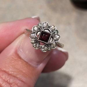 Pu&ograve; includere: Un anello color argento con una pietra centrale rosso scuro a taglio quadrato, circondata da un'aureola a forma di fiore composta da piccole pietre chiare a taglio rotondo. La montatura ha una finitura testurizzata e scura, con un gambo liscio.