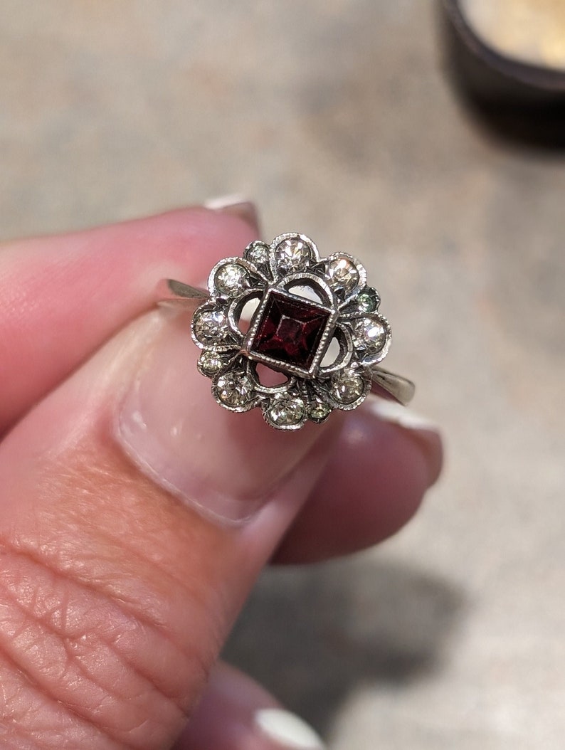 Pu&ograve; includere: Un anello color argento con una pietra centrale rosso scuro a taglio quadrato, circondata da un'aureola a forma di fiore composta da piccole pietre chiare a taglio rotondo. La montatura ha una finitura testurizzata e scura, con un gambo liscio.