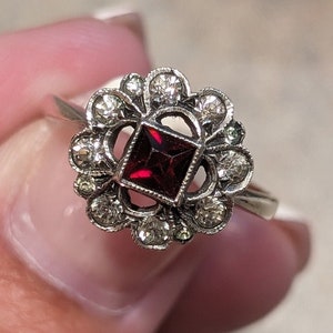 Pu&ograve; includere: Un anello color argento con una pietra preziosa rosso scuro a taglio quadrato al centro. La pietra &egrave; circondata da una montatura decorativa a forma di fiore, ornata da diverse piccole pietre chiare a taglio rotondo disposte a cerchio.