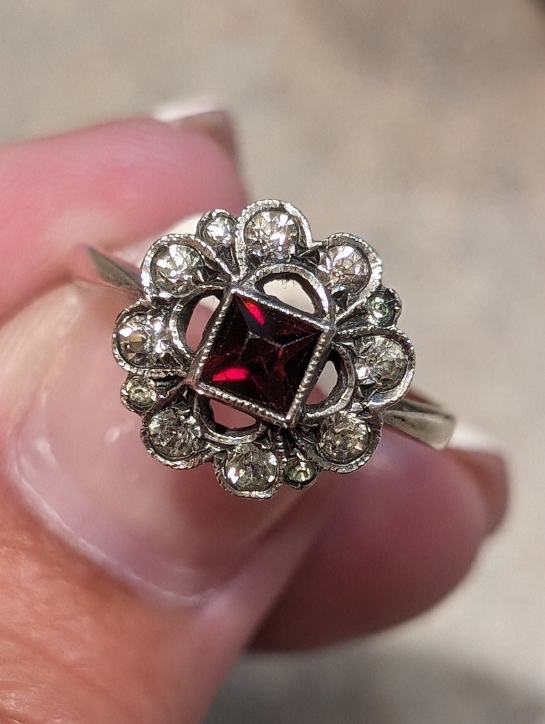Pu&ograve; includere: Un anello color argento con una pietra preziosa rosso scuro a taglio quadrato al centro. La pietra &egrave; circondata da una montatura decorativa a forma di fiore, ornata da diverse piccole pietre chiare a taglio rotondo disposte a cerchio.