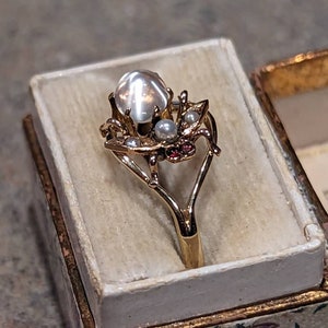 K&ouml;nnte beinhalten: Goldfarbener Ring mit einem gro&szlig;en, schillernden, runden Edelstein in der Mitte, umgeben von kleineren Perlen und roten Akzenten. Der Ring wird in einer Schatulle mit floralem Muster und cremefarbenem Innenraum pr&auml;sentiert.