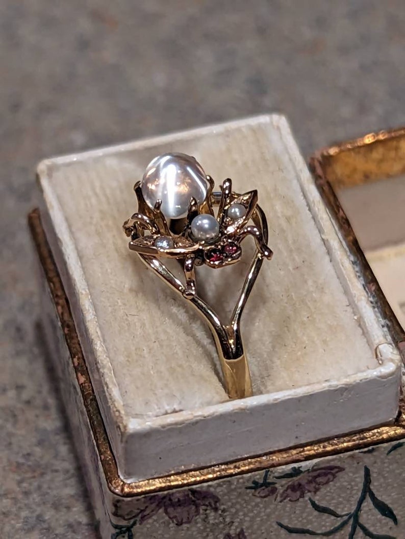 K&ouml;nnte beinhalten: Goldfarbener Ring mit einem gro&szlig;en, schillernden, runden Edelstein in der Mitte, umgeben von kleineren Perlen und roten Akzenten. Der Ring wird in einer Schatulle mit floralem Muster und cremefarbenem Innenraum pr&auml;sentiert.