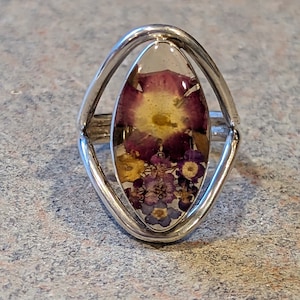 May include: A silver ring with a clear oval-shaped stone containing pressed purple and yellow flowers.