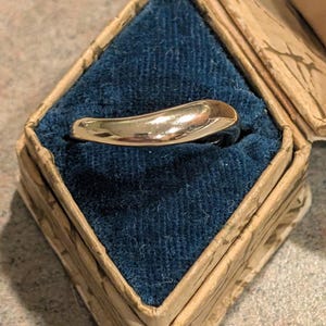 Könnte beinhalten: Ein goldfarbener Ring mit glattem, geschwungenem Design, präsentiert in einer Vintage-Box in Diamantenform mit blauem Samtinterieur. Die Box hat eine abgenutzte, gemusterte Außenseite.