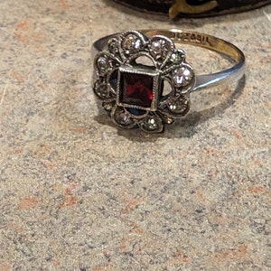 Pu&ograve; includere: Un anello color argento con una pietra preziosa rosso scuro a taglio quadrato al centro, circondata da un alone di piccole pietre rotonde sfaccettate. La fascia in metallo &egrave; sottile e lucida, con un marchio visibile sulla superficie interna.
