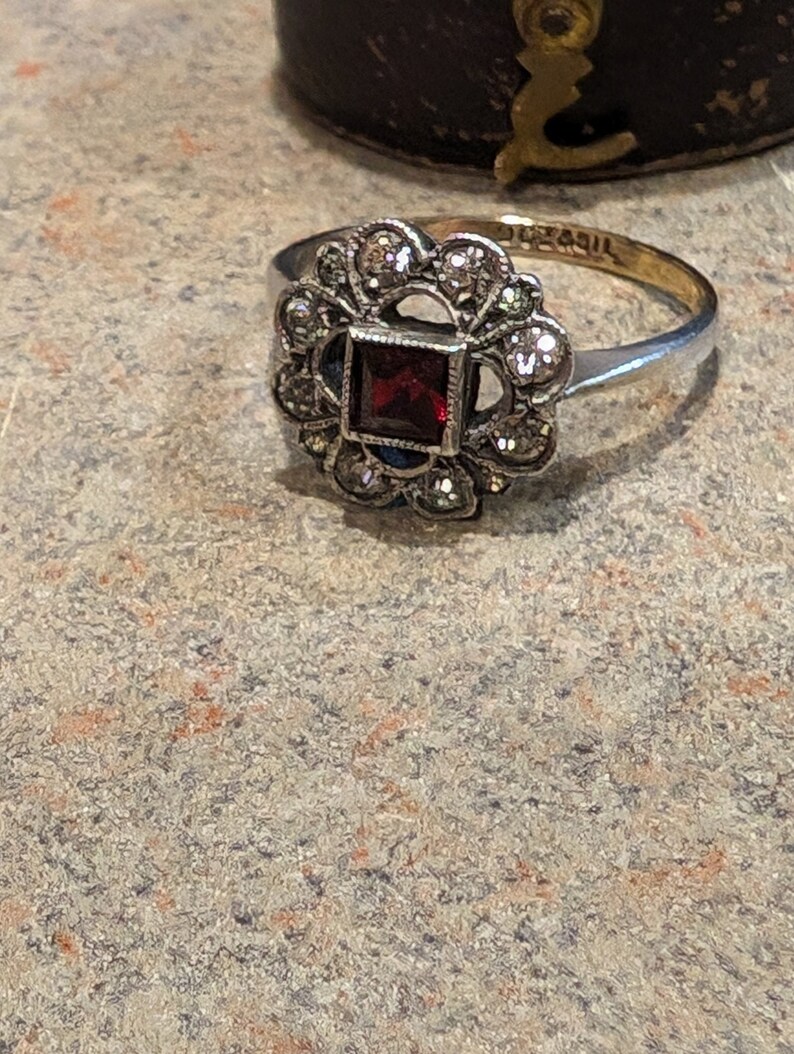 Pu&ograve; includere: Un anello color argento con una pietra preziosa rosso scuro a taglio quadrato al centro, circondata da un alone di piccole pietre rotonde sfaccettate. La fascia in metallo &egrave; sottile e lucida, con un marchio visibile sulla superficie interna.
