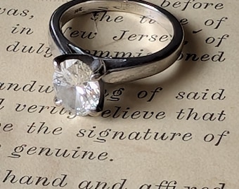 Op de afbeelding: Een zilveren ring met een grote, heldere, vierkant geslepen edelsteen. De ring is geplaatst tegen een achtergrond van verouderd papier met vervaagde tekst, wat een vintage esthetiek creëert. De band van de ring is glad en gepolijst.