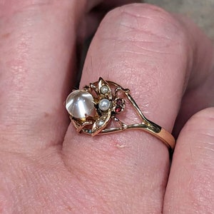 K&ouml;nnte beinhalten: Ein goldener Ring mit einem gro&szlig;en, runden, opalisierenden Stein. Der Ring ist mit kleinen Perlen, roten Edelsteinen und blattartigen Details verziert. Das Band hat ein aufwendiges, offenes Design.