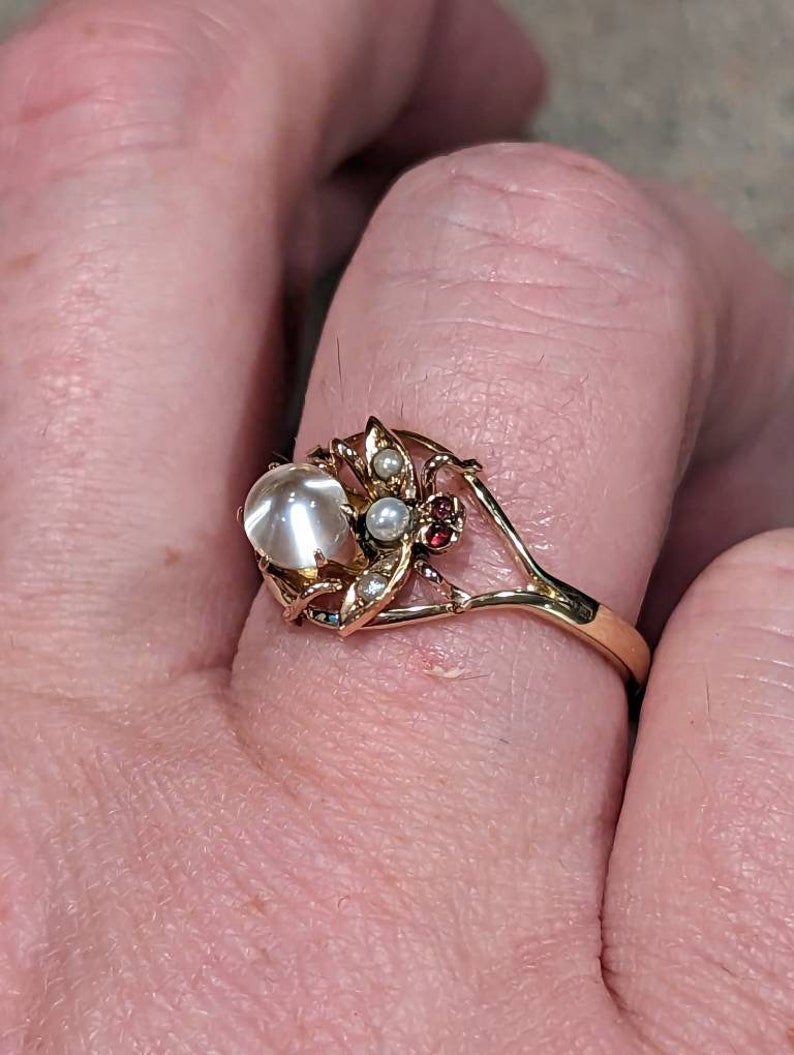 K&ouml;nnte beinhalten: Ein goldener Ring mit einem gro&szlig;en, runden, opalisierenden Stein. Der Ring ist mit kleinen Perlen, roten Edelsteinen und blattartigen Details verziert. Das Band hat ein aufwendiges, offenes Design.
