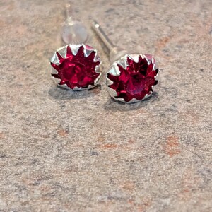 May include: Stud earrings with a bright red gemstone in a silver-coloured, starburst frame. The earrings are shown on a textured, neutral surface, highlighting their sparkling design. The earrings are suitable for everyday wear.