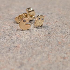May include: Gold-colored stud earrings featuring a Pac-Man and ghost design. The Pac-Man has a clear gem in the center, and the ghost has two clear gems for eyes. The earrings are on a speckled, neutral-toned surface.
