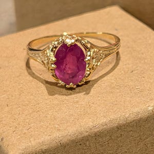 May include: A gold ring featuring an oval, deep pink gemstone. The ring has an ornate, filigree design around the gemstone and on the band. The ring is set on a brown surface.