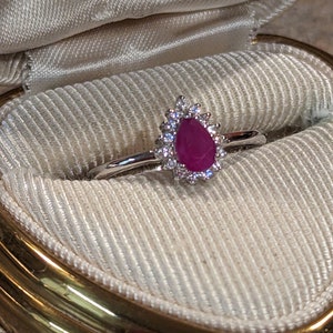 May include: A silver-toned ring featuring a pear-shaped, deep pink gemstone at its centre. The central stone is surrounded by a halo of small, clear, sparkling stones set in a metallic frame, mounted on a thin, polished silver-toned band.