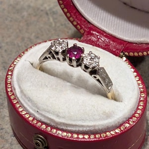 May include: A three-stone ring with a central ruby and two diamond side stones, set in a gold and silver band. The ring is displayed in a red jewelry box with a white interior.