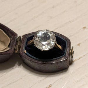 May include: A clear, faceted gemstone ring set in a gold band. The ring is displayed in a small, dark brown velvet ring box.