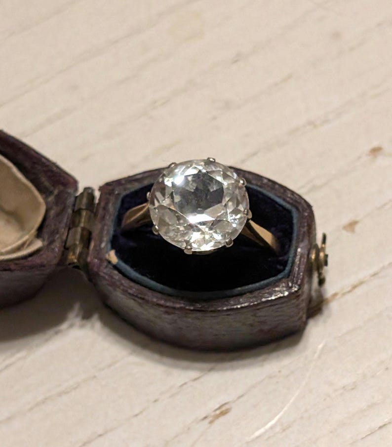 May include: A clear, faceted gemstone ring set in a gold band. The ring is displayed in a small, dark brown velvet ring box.
