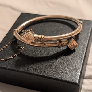 May include: A gold-toned hinged bangle bracelet with decorative square accents and a delicate chain. The bracelet rests on a black box, showcasing its intricate design and vintage appeal. The square accents feature an engraved leaf pattern.