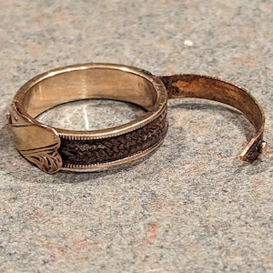 May include: A gold-toned ring with a decorative design on the band. The ring is open on one side and has a hinge.