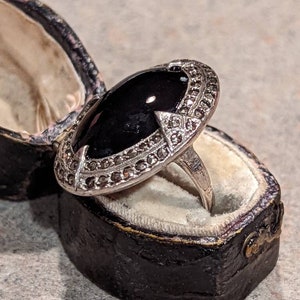 Op de afbeelding: Een ovale zwarte edelsteenring met een zilverkleurige zetting, gepresenteerd in een vintage zwarte sieradendoos. De zetting van de ring is versierd met kleine, sprankelende accenten. De doos heeft een crèmekleurig interieur.